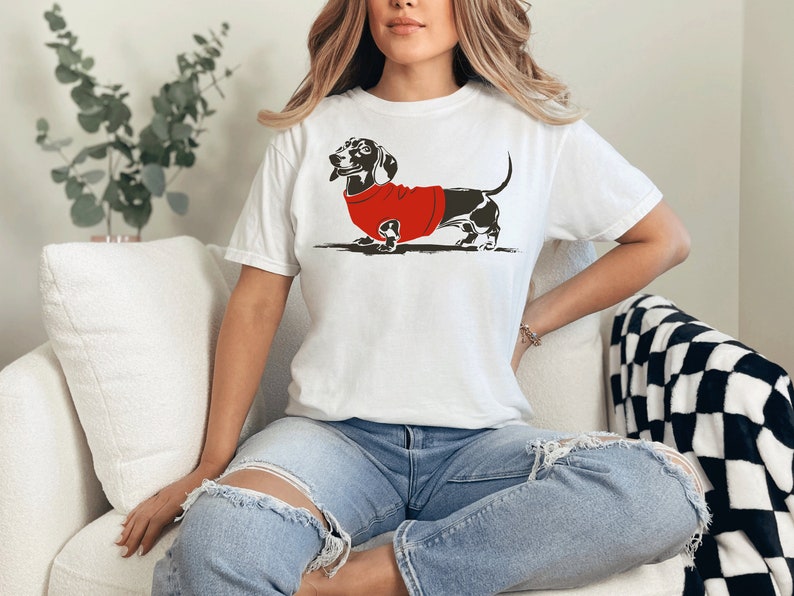May include: A white t-shirt featuring a black and white illustration of a dachshund wearing a red jumper.