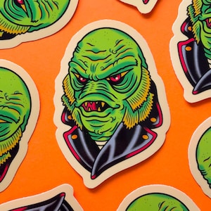 May include: A green and black sticker featuring a cartoon illustration of the Creature from the Black Lagoon wearing a leather jacket. The creature has red eyes, sharp teeth, and gills.