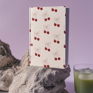 Op de afbeelding: Een crèmekleurig notitieboek met een patroon van rode kersen en strikken. Het notitieboek staat op een lichtgrijze rotsformatie, met een glas groene vloeistof met ijs op de achtergrond. De achtergrond is lichtpaars.