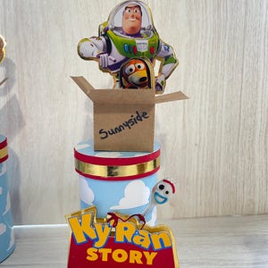 May include: A blue and white cylinder with a gold trim and a red and gold banner that says "Toy Story" with a yellow and red banner that says "Ky Ran" on top. A brown box with the word "Sunnyside" written on it sits on top of the cylinder. A cardboard cutout of Buzz Lightyear and Woody from Toy Story is on top of the box.