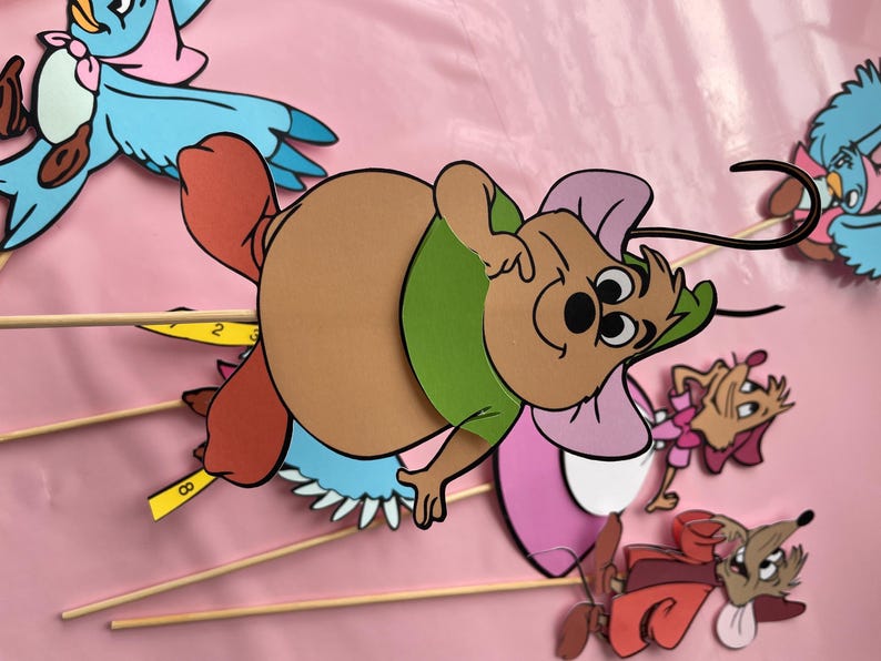 May include: A set of five cartoon character cutouts on sticks. The characters include a blue bird, a brown mouse with a green shirt, a pink mouse with a white dress, a brown mouse with a red dress, and a brown mouse with a pink dress.