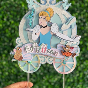 May include: A blue and silver Cinderella themed cake topper with a glitter finish. The topper features Cinderella in her blue dress and glass slipper, a carriage, two mice, and the name "Allison".