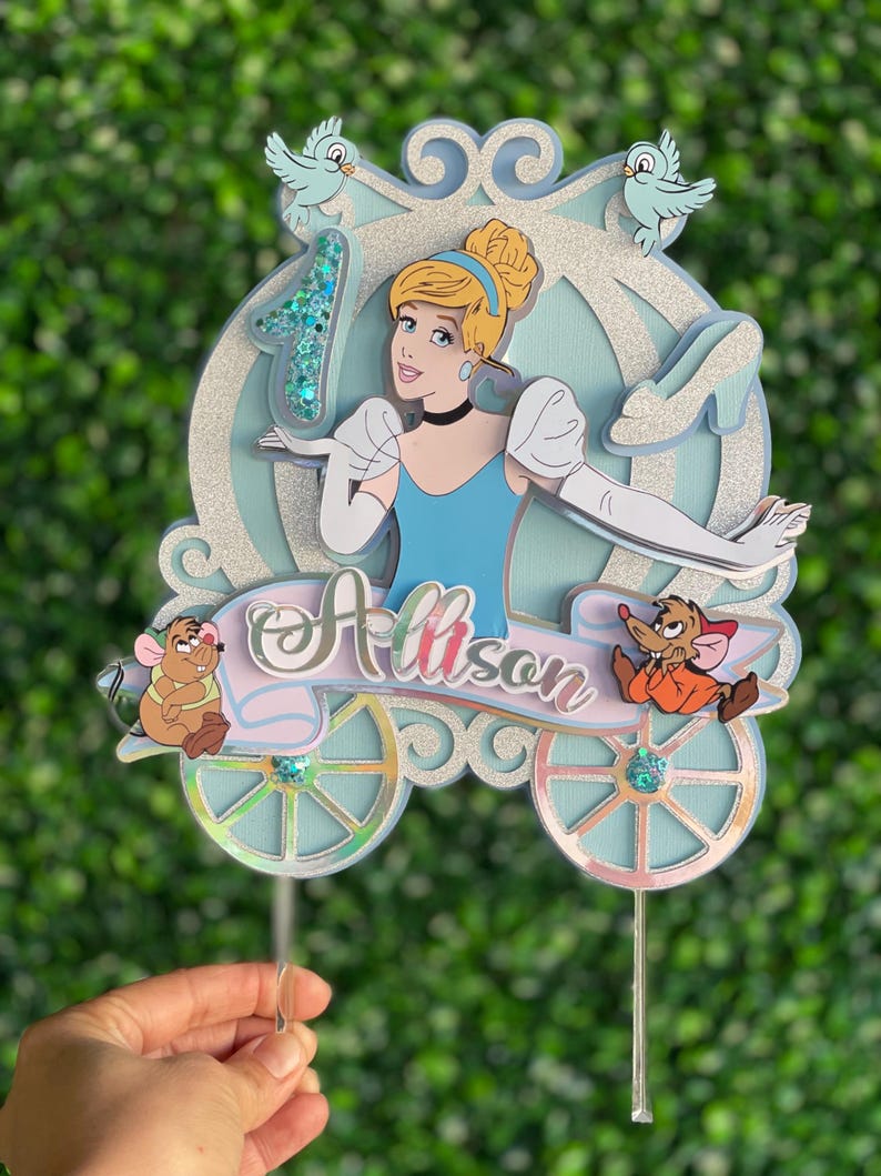 May include: A blue and silver Cinderella themed cake topper with a glitter finish. The topper features Cinderella in her blue dress and glass slipper, a carriage, two mice, and the name "Allison".