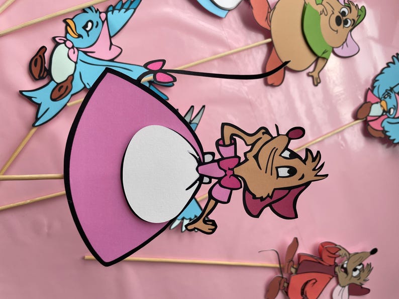May include: A set of five paper cutouts of characters from the Disney movie Cinderella. The characters include Cinderella, Gus, Jaq, the mice, and Lucifer, the cat. The cutouts are on sticks and are ready to be used as party decorations.
