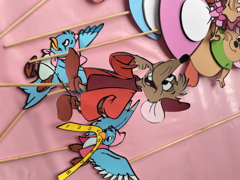 May include: A set of six colorful paper cutouts of cartoon characters on sticks. The characters include a mouse wearing a red coat, a blue bird, and a pink and white bird. The characters are perfect for a children's party or for use as props in a play.