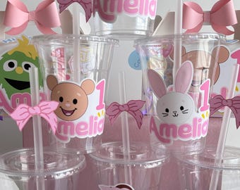 May include: Six clear plastic cups with pink and white personalized labels. Each cup has a different character on it, including a girl, a bear, a bunny, a green monster, and an orange monster. The labels all say "Amelia" and "1".