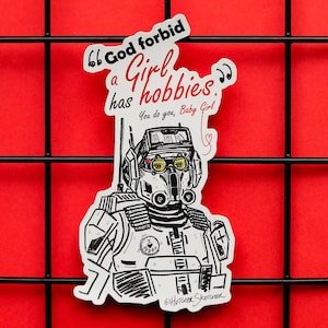 May include: A white sticker featuring a black and white illustration of a robot-like character. The sticker includes red and black text: "God forbid a Girl has hobbies. You do you, Baby Girl."
