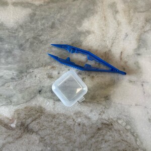 May include: A blue plastic tweezers and a clear plastic container. The tweezers have a serrated grip and are designed for picking up small objects.