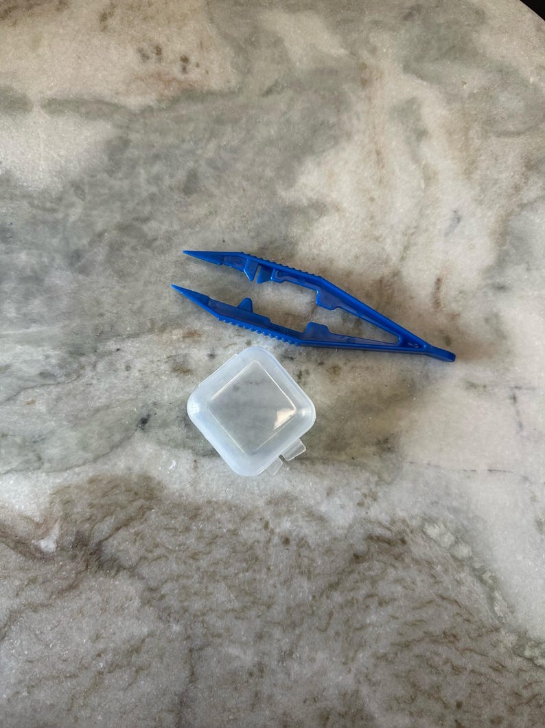 May include: A blue plastic tweezers and a clear plastic container. The tweezers have a serrated grip and are designed for picking up small objects.