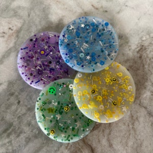 May include: Four round coasters with different designs. Each coaster has a translucent background with embedded glitter and small plastic shapes. The designs include blue glitter with white stars, purple glitter with black smiley faces, yellow glitter with yellow smiley faces, and green glitter with green stars.