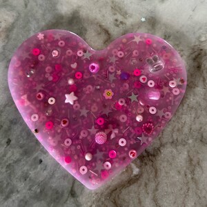 May include: A pink heart-shaped resin tray filled with various pink, white, and silver glitter and beads.