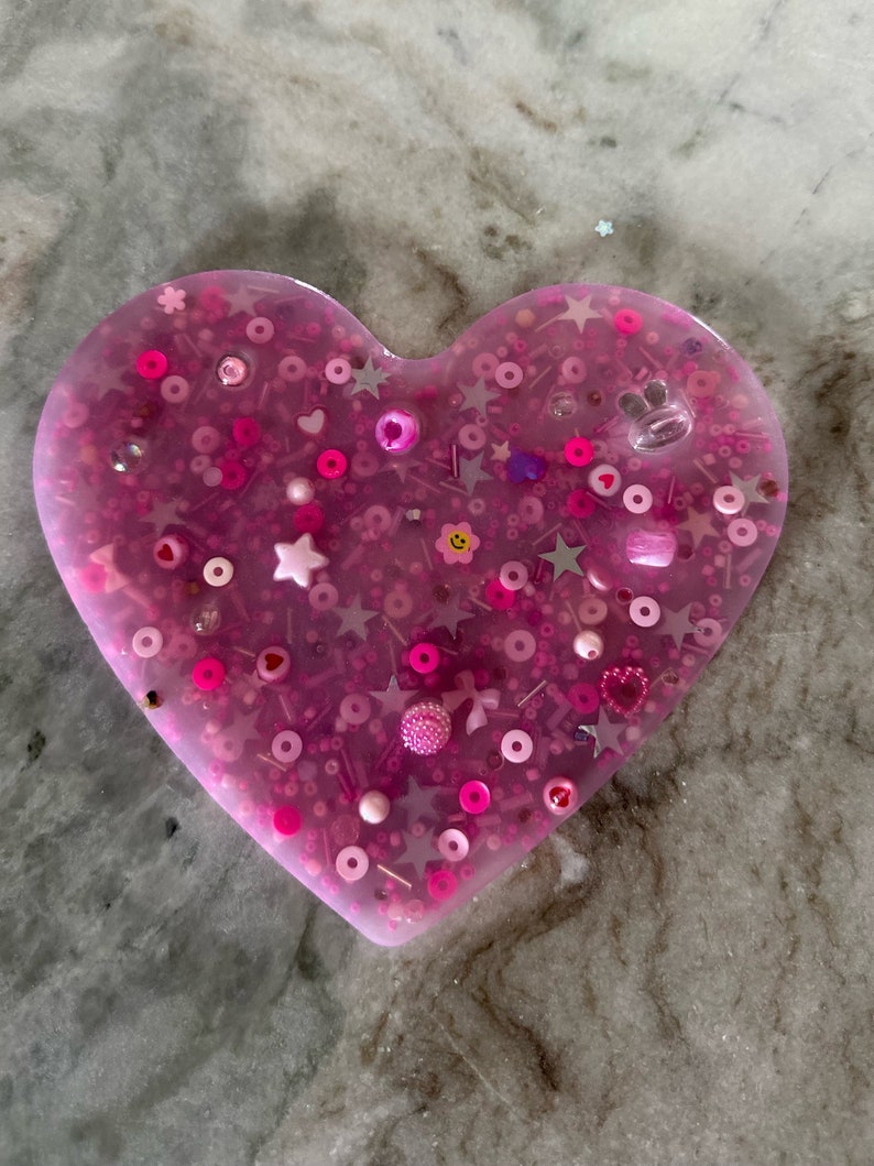 May include: A pink heart-shaped resin tray filled with various pink, white, and silver glitter and beads.