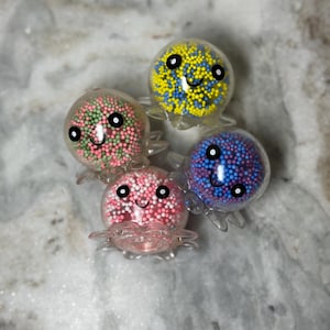 May include: Four clear plastic octopus figurines filled with colorful sprinkles. Each octopus has a smiling face with black eyes and a pink, blue, green, or yellow body.