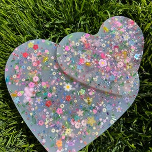 May include: Two heart-shaped resin coasters with a light blue background and a variety of colorful beads, flowers, and other small decorative elements embedded in the resin.