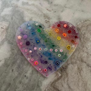 May include: A heart-shaped resin coaster with a rainbow gradient and embedded glitter, beads, and charms. The name "MAGGIE" is spelled out in colorful beads.