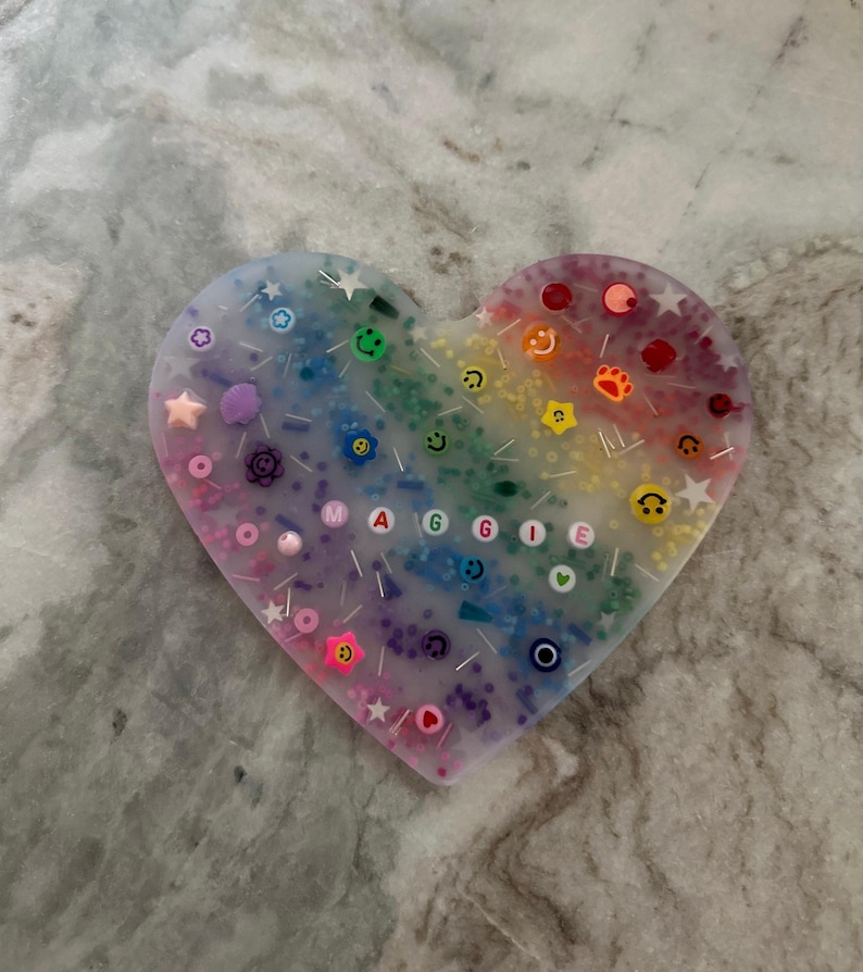 May include: A heart-shaped resin coaster with a rainbow gradient and embedded glitter, beads, and charms. The name "MAGGIE" is spelled out in colorful beads.