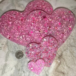 May include: Set of four pink heart-shaped resin coasters with glitter and various embellishments. The coasters are arranged in a pyramid shape, with the largest coaster at the bottom and the smallest coaster at the top.