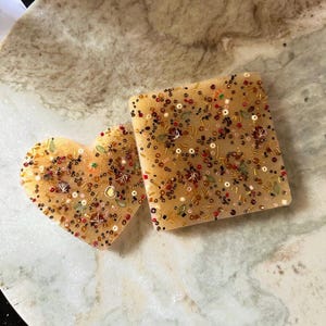 Könnte beinhalten: Zwei dekorative Seifen, eine herzförmige und eine quadratische, werden gezeigt. Beide sind hellbernsteinfarben und stark mit kleinen Perlen und dekorativen Elementen in Rot-, Gold- und Grüntönen verziert. Die Seifen liegen auf einer Marmoroberfläche.