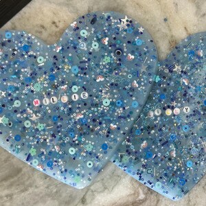 May include: Two blue resin heart-shaped ornaments with glitter and colorful beads. The larger heart has the name "Millie" spelled out in white beads.