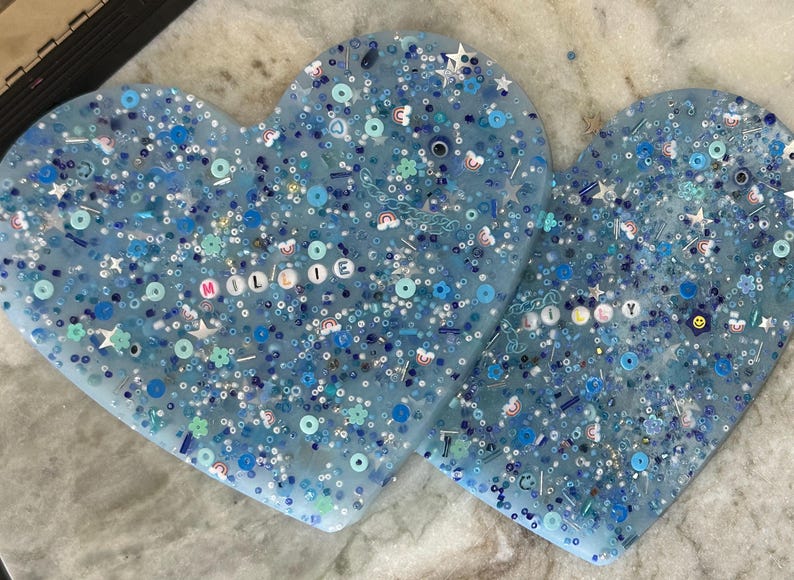 May include: Two blue resin heart-shaped ornaments with glitter and colorful beads. The larger heart has the name "Millie" spelled out in white beads.