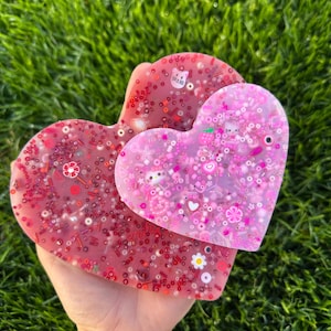 Könnte beinhalten: Zwei herzförmige Untersetzer aus Harz mit eingebettetem Glitzer und bunten Verzierungen. Der größere Untersetzer ist pink mit weißem Glitzer und der kleinere Untersetzer ist rot mit pinkem Glitzer.