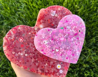 May include: Two heart-shaped resin coasters with embedded glitter and colorful embellishments. The larger coaster is pink with white glitter and the smaller coaster is red with pink glitter.
