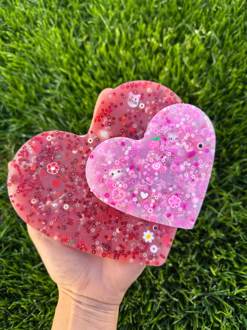 May include: Two heart-shaped resin coasters with embedded glitter and colorful embellishments. The larger coaster is pink with white glitter and the smaller coaster is red with pink glitter.