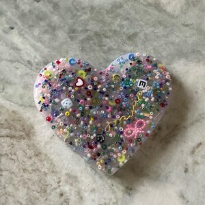 May include: A heart-shaped resin ornament filled with colorful beads, sequins, and small charms. The ornament is translucent and has a light pink hue.
