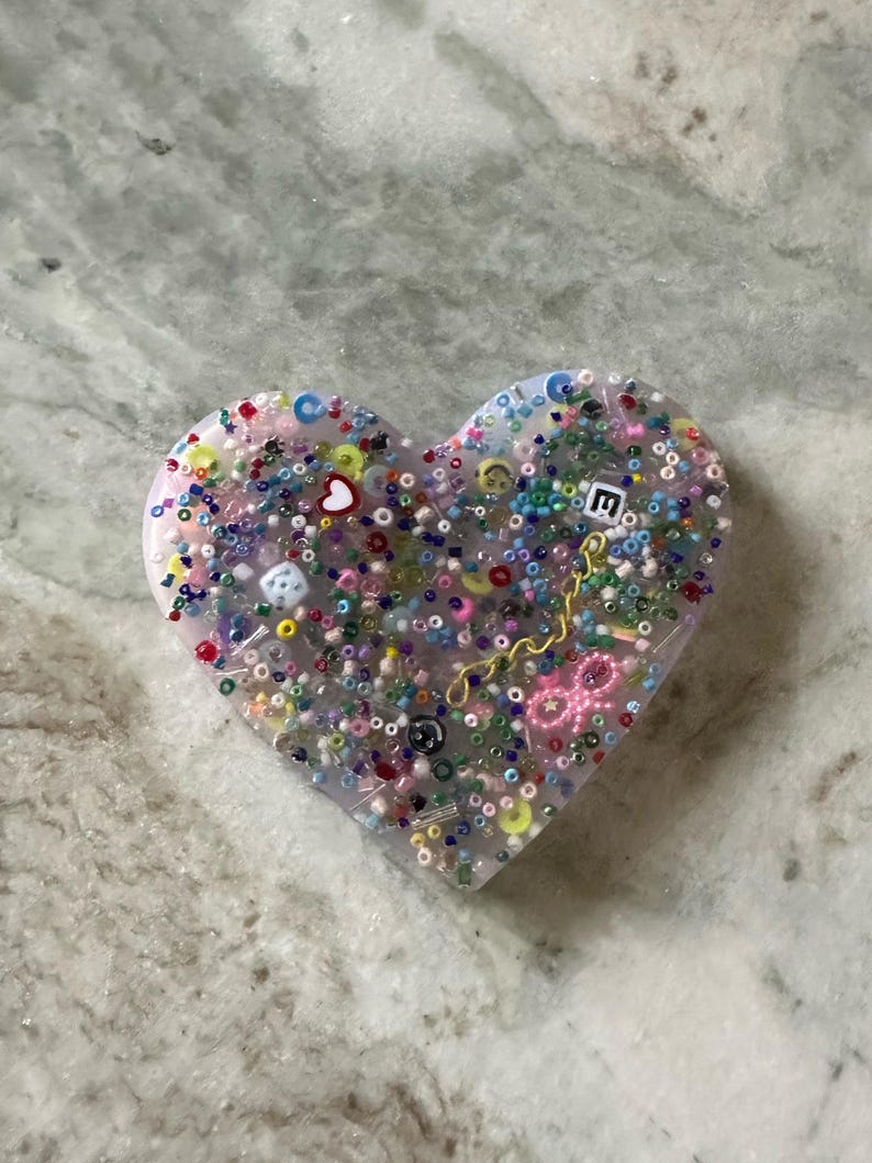 May include: A heart-shaped resin ornament filled with colorful beads, sequins, and small charms. The ornament is translucent and has a light pink hue.
