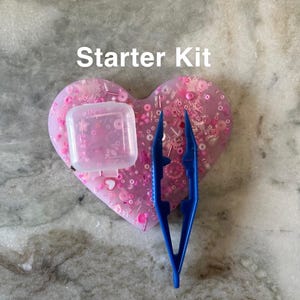 May include: A pink heart-shaped tray filled with pink and white beads, sequins, and other embellishments. A small clear plastic container and blue tweezers are also included. The text "Starter Kit" is written above the tray.