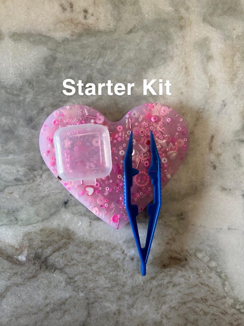 May include: A pink heart-shaped tray filled with pink and white beads, sequins, and other embellishments. A small clear plastic container and blue tweezers are also included. The text "Starter Kit" is written above the tray.