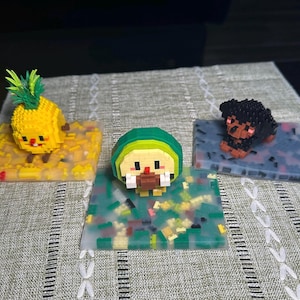 May include: Three miniature figurines made of colorful plastic building blocks. The figurines are a pineapple, a green avocado, and a black dog. Each figurine is displayed on a small base of assorted plastic blocks.