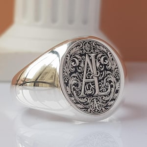 Puede incluir: Un anillo de sello de plata con un diseño grabado en negro que presenta las letras "A L" rodeadas de volutas ornamentadas.