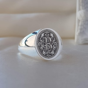 May include: A polished silver signet ring featuring an oval face with a deeply engraved, ornate letter B centered within intricate scrollwork and filigree patterns. The band is smooth and reflective, showcasing a classic, solid metal construction.