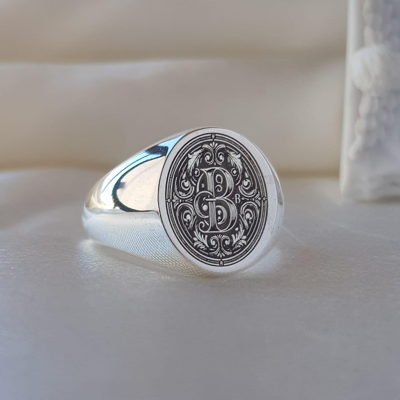 May include: A polished silver signet ring featuring an oval face with a deeply engraved, ornate letter B centered within intricate scrollwork and filigree patterns. The band is smooth and reflective, showcasing a classic, solid metal construction.