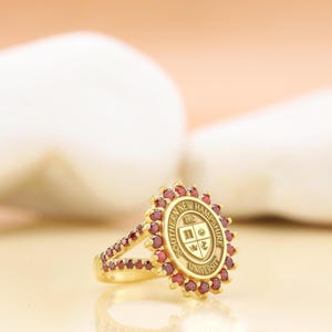 May include: A gold ring featuring a central oval emblem with the text Southern New Hampshire University and the year 1932. The emblem is surrounded by a halo of small, round, dark red stones, which also extend down the split shank of the ring.