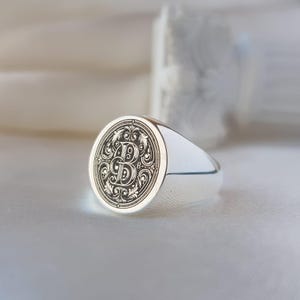 May include: A polished silver signet ring featuring a circular face with an intricate, dark engraved letter B surrounded by ornate, symmetrical scrollwork patterns. The band is smooth and metallic, reflecting light across its curved surface.