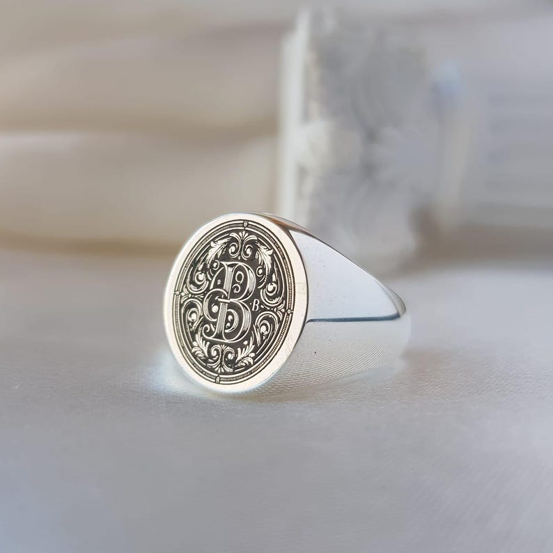 May include: A polished silver signet ring featuring a circular face with an intricate, dark engraved letter B surrounded by ornate, symmetrical scrollwork patterns. The band is smooth and metallic, reflecting light across its curved surface.