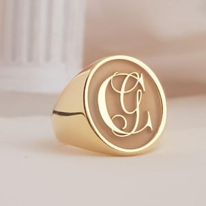 May include: A gold signet ring with a polished finish. The ring features a circular face with the capital letter "G" in an elegant script. The ring is set against a soft, neutral background, highlighting the ring's design.