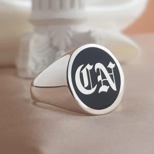 May include: A silver signet ring featuring a circular face with a black inlay. The center of the ring displays the letters CN in a white, gothic-style font. The band is polished silver with a smooth, reflective finish and a tapered design.