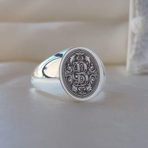 May include: A polished silver signet ring featuring an oval face with a detailed black engraved design. The center of the oval displays a large, ornate letter B surrounded by intricate scrollwork and filigree patterns within a decorative border.
