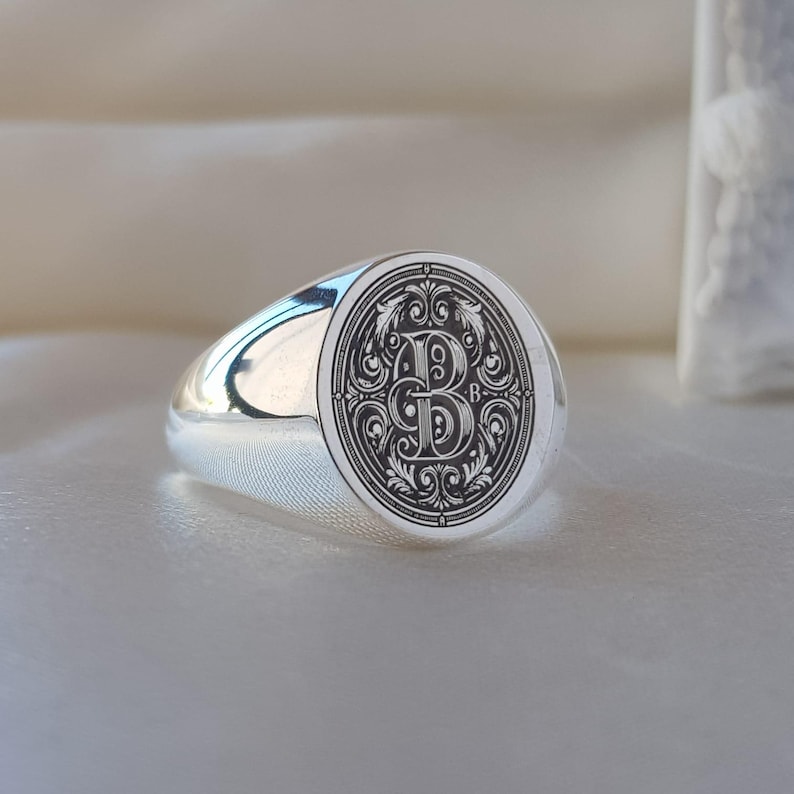 May include: A polished silver signet ring featuring an oval face with a detailed black engraved design. The center of the oval displays a large, ornate letter B surrounded by intricate scrollwork and filigree patterns within a decorative border.