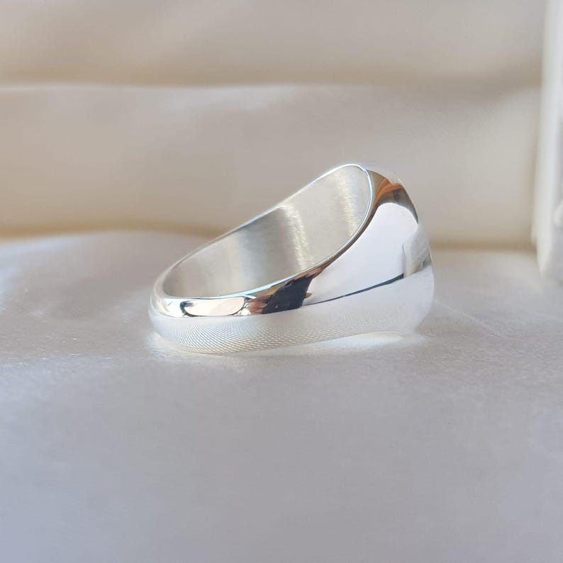 May include: A polished silver signet ring with a smooth, reflective surface and a rounded band. The ring features a solid, thick construction with a high-shine metallic finish that catches the light, showcasing a clean, minimalist design.