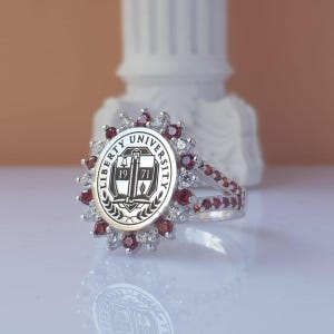 May include: Silver ring featuring the Liberty University seal. The ring has a round center with the university logo and the text "LIBERTY UNIVERSITY 19 71". The ring is adorned with red and white gemstones.