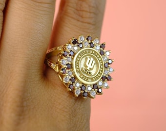 May include: A gold-colored ring featuring the University of Indianapolis logo. The ring has a circular center with the university name and a stylized logo. It is surrounded by alternating red and white gemstones, with the year 2023 inscribed.