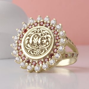 May include: A gold ring featuring a circular center plate engraved with the letters CG surrounded by intricate scrollwork. The center is framed by a double halo of alternating clear white stones and deep red stones in a sunburst pattern.