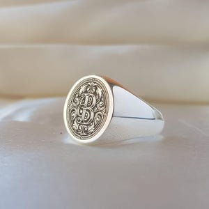 May include: A polished silver signet ring featuring a circular face with an intricate, engraved letter B surrounded by ornate, swirling filigree patterns. The band is smooth and reflective, showcasing a classic design with a detailed monogram.