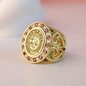 May include: A gold ring with a large circular centerpiece featuring the words "Coroner of the Year" and a seal. The ring is adorned with red and white gemstones.
