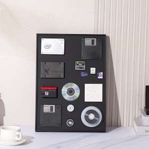 Può includere: Una lavagna nera incorniciata con vari componenti hardware per computer, tra cui floppy disk, dischi rigidi e schede di memoria. La lavagna è montata su una parete bianca. Una piccola statuetta di un subacqueo è in primo piano.