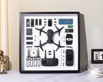 May include: A black drone with its parts laid out in a white frame. The drone has four propellers and a camera. The parts include the battery, charger, and remote control. The frame is made of wood and has a black border.